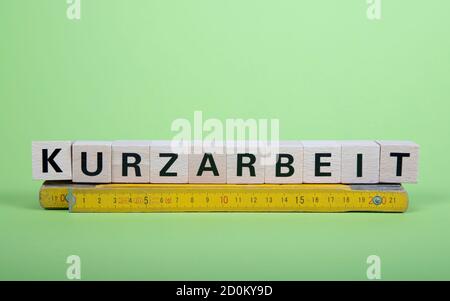 wooden blocks with german word for short time work, Kurzarbeit, above a folding rule, green background Stock Photo