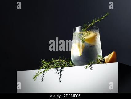 Alcohol drink (gin tonic cocktail) with lemon, juniper branch, and ice on a dark reflective background, copy space. Iced cocktail drink with lemon. Stock Photo