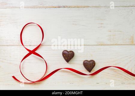 Red gift celebration ribbon waved in shape of number 8 (eight) and chocolate in heart shape on white wooden background. Greeting card with copy space Stock Photo