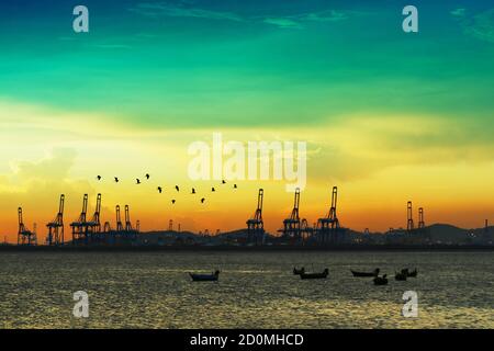 birds flying over cranes on seaport and sunset colorful sky and many fishing boats are parking on sea Stock Photo
