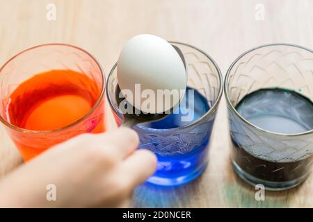 Transparent glass cups with colored water. The process of coloring eggs. A child hands put in a white egg with a spoon. Stock Photo