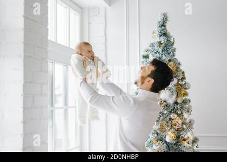 A young dad holds him in his arms while lifting him up near the Christmas tree in the living room in the house Stock Photo