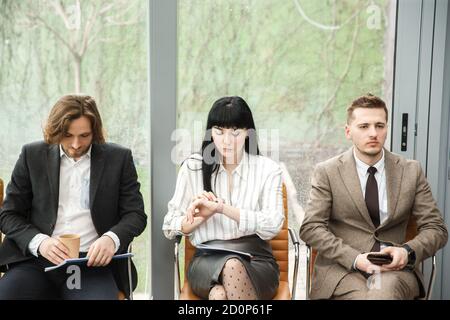 Three office workers are waitig for an appointment. The woman in the middle is looking at her watch. Stock Photo