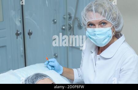 Portrait of  middle aged woman doctor cosmetician in white lab coat, mask and sterile gloves makes injection of Botox into the forehead of a young wom Stock Photo