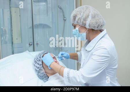 Portrait of  middle aged woman doctor cosmetician in white lab coat, mask and sterile gloves makes injection of Botox into the forehead of a young wom Stock Photo