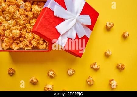 Red gift box with white bow full of tasty caramel popcorn on yellow background. Stock Photo