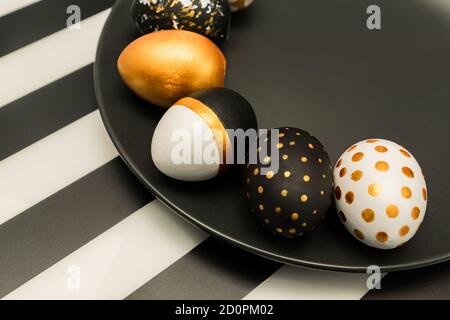 Close up of black, white and golden decorated eeaster eggs on black plate on striped background. Trendy holiday background. Stock Photo