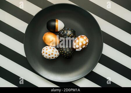 Black and white striped background with white, black and golden decorated eggs on black plate. Trendy easter flat lay. Stock Photo