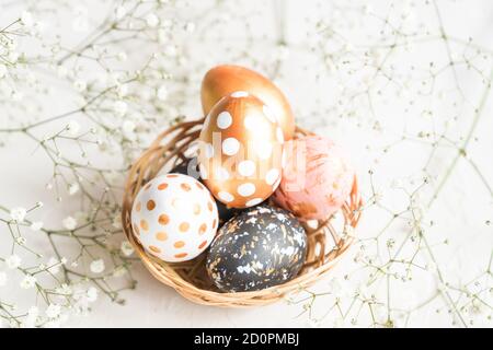 Close up of black, white and golden decorated eeaster eggs in wicker basket decorated with gypsophila on white background. Stock Photo