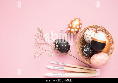 Top view of wooden painted eggs in gold, black and rose colors with branch of gypsophila and paint brushes on pink background. Happy easter background Stock Photo