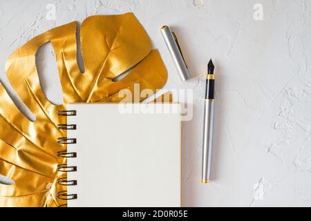 Stylish home office desk with  paper notepad, golden pen and tropical monstera leaf. Flat lay, top view, copy space. Feminine workspace concept. Stock Photo