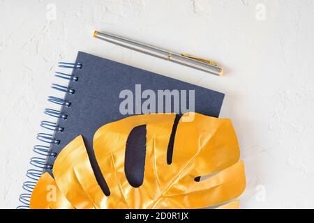 Stylish home office desk with  paper notepad, golden pen and tropical monstera leaf. Flat lay, top view, copy space. Feminine workspace concept. Stock Photo