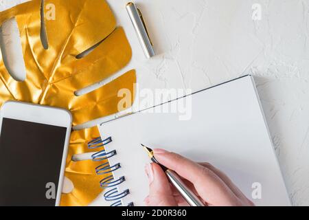 Top view of woman desktop. Golden tropical monstera leaf, blank notepad, white smartphone and pen on white concrete background. Stylish minimal layout Stock Photo