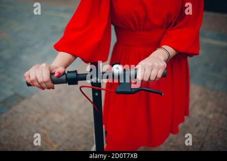 Young woman holds steering wheel of electric scooter in red dress at the city Stock Photo
