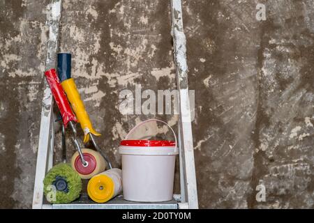 Colored construction rollers with a bucket stand on a stepladder on the background of an untreated concrete wall with paint remnants. Background. Stock Photo
