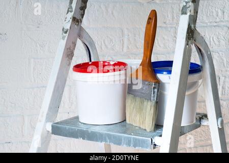 A paintbrush and buckets with red and blue lids close up stand on a metal stepladder against a white wall decorated with bricks. Background. Stock Photo