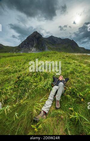 Woman lies pleasurably in a meadow in front of threatening mountain range, Skagsanden, Flakstad, Lofoten, Nordland, Norway, Europe