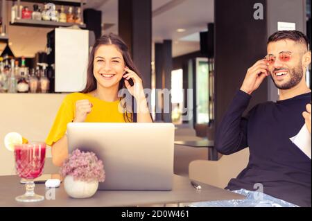 Two young  modern business guys sitting in a bar, using laptop, looking each other, smiling and chatting - Active people watching a funny streaming mo Stock Photo