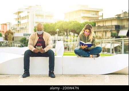Boy working on the tablet whilst  a girl reading her book with protective mask sitting on the bench in the park - Couple relaxing at seaside at sunset Stock Photo