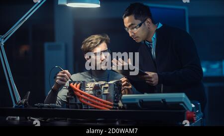 Two Professional Automotive Engineers with a Tablet Computer and Inspection Tools are Having a Conversation While Testing an Electric Engine in a High Stock Photo