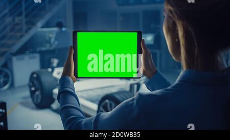 Female Engineer Holds a Tablet Computer with Green Screen Mock Up and Looks at Electric Car Chassis Prototype with Wheels, Batteries and Engine Stock Photo