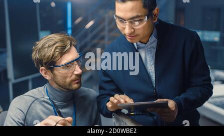 Two Professional Automotive Engineers with a Tablet Computer and Inspection Tools are Having a Conversation While Testing an Electric Engine in a High Stock Photo