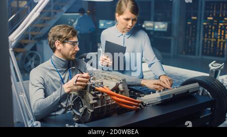 Male and Female Automotive Engineers with a Tablet Computer and Inspection Tools are Having a Conversation While Testing an Electric Engine in a High Stock Photo