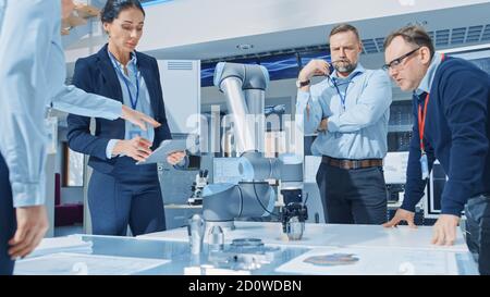 Diverse Team of Industrial Robotics Engineers Gathered Around Table With Robot Arm, They Manipulate and Program it to Pick Up and Move Metal Component Stock Photo