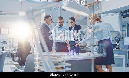 Diverse Team of Industrial Robotics Engineers Gathered Around Table With Robot Arm, They Use Tablet Computer to Manipulate and Program it to Pick Up Stock Photo