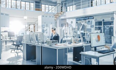 Female Chef Engineer Working on Computer, Programming Robot Arm in Bright Modern Industrial Robotics Technology and Design Office. Working in Research Stock Photo