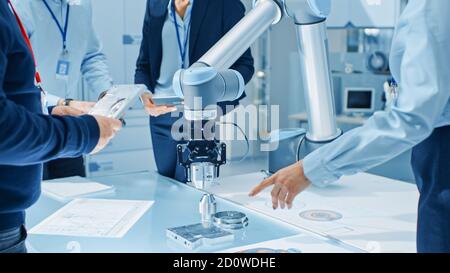 Team of Industrial Robotics Engineers Gathered Around Table With Robot Arm, They Use Tablet Computer to Manipulate and Program it to Pick Up and Move Stock Photo