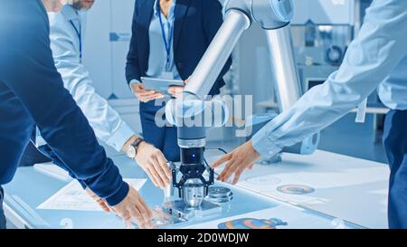 Team of Industrial Robotics Engineers Gathered Around Table With Robot Arm, They Use Tablet Computer to Manipulate and Program it to Pick Up and Move Stock Photo