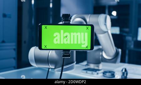 Robot Arm Holding Green Mock-up Screen Smartphone in Landscape Mode. Industrial Robotic Manipulator End Effector Holds Mobile Phone with Chroma Key Stock Photo