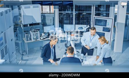 Diverse Team of Industrial Robotics Engineers Gathered Around Table With Robot Arm, They Manipulate and Program it to Pick Up and Move Metal Component Stock Photo