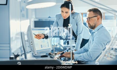 Female Chief Engineer Talks with Male Electronics Specialist, Explaining Things, He Works on Computer and Agrees with Her. Modern and Bright Office Stock Photo