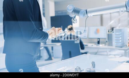 Stylish Female Industrial Robotics Engineer Uses Tablet Computer with CAD Software and to Manipulate and Program Robot Arm for Lifting, Moving Objects Stock Photo