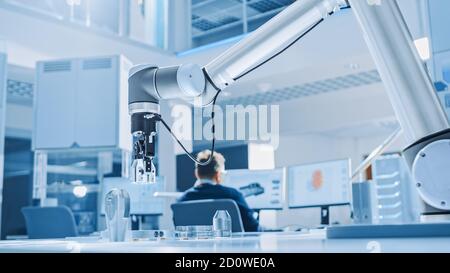 Modern Robot Arm Picks up, Lifts and Moves Metal Component as Programmed. In the Background Industrial Robotics Engineers Working on Software Stock Photo
