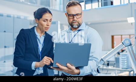 Female Chief Engineer Talks with Male Electronics Specialist, Explaining Things, He Holds on Laptop Computer. Modern and Bright Office with Stylish Stock Photo