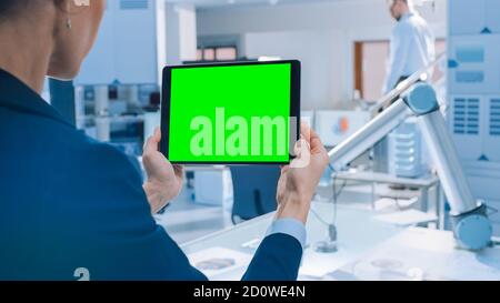 Engineer Holds Green Mock-up Screen Digital Tablet Computer. In the Background Team of Industrial Engineers Working with Functional Robot Arm in Stock Photo