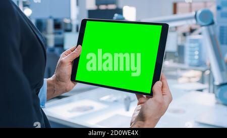 Engineer Holds Green Mock-up Screen Digital Tablet Computer. In the Background Team of Industrial Engineers Working with Functional Robot Arm in Stock Photo