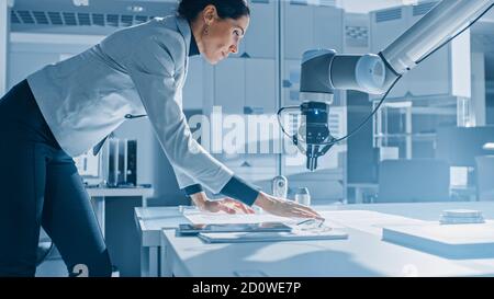 Stylish Female Robotics Engineer Leans on the Table Works with Blueprints, Documents and Tablet Computer, She's Programming Robot Arm Movements Stock Photo