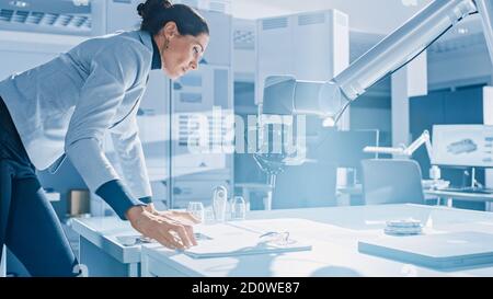 Stylish Female Robotics Engineer Leans on the Table Works with Blueprints, Documents and Tablet Computer, She's Programming Robot Arm Movements Stock Photo