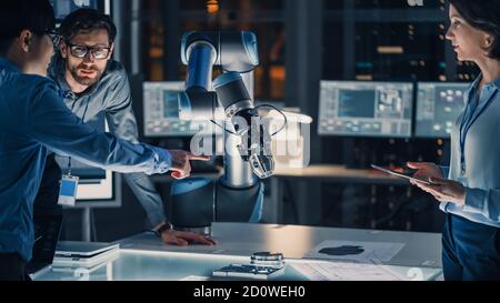 Diverse Team of Engineers with Laptop and a Tablet Analyse and Discuss How a Futuristic Robotic Arm Works and Moves a Metal Object. They are in a High Stock Photo