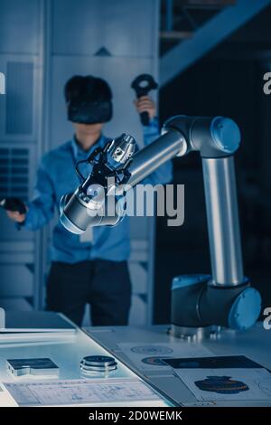 Vertical Shot of Professional Development Engineer in Blue Shirt Controlling Futuristic Robotic Arm with Virtual Reality Headset and Joysticks in a Stock Photo