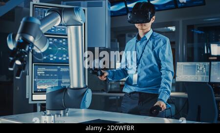 Professional Japanese Development Engineer in Blue Shirt is Controlling a Futuristic Robotic Arm with a Virtual Reality Headset and Joysticks in a Stock Photo