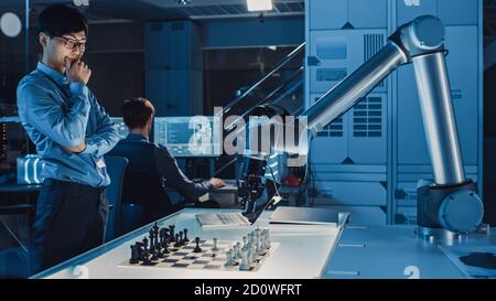Professional Japanese Development Engineer is Testing an Artificial Intelligence Interface by Playing Chess with a Futuristic Robotic Arm. They are in Stock Photo