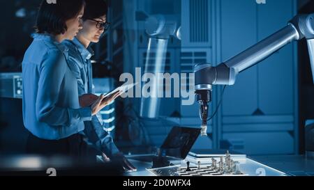 Two Development Engineers are Discussing and Testing an Artificial Intelligence Interface by Playing Chess with a Futuristic Robotic Arm. They are in Stock Photo
