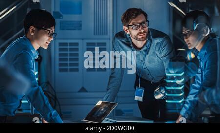 Diverse Team of Engineers with Laptop and a Tablet Analyse and Discuss How a Futuristic Robotic Arm Works and Moves a Metal Object. They are in a High Stock Photo
