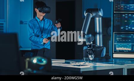 Professional Japanese Development Engineer in Blue Shirt is Controlling a Futuristic Robotic Arm with a Virtual Reality Headset and Joysticks in a Stock Photo