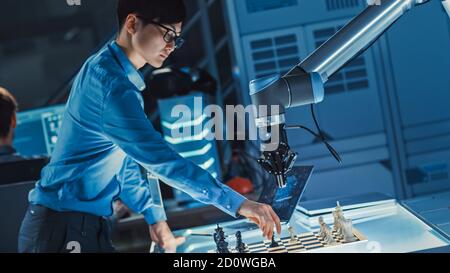 Professional Japanese Development Engineer is Testing an Artificial Intelligence Interface by Playing Chess with a Futuristic Robotic Arm. They are in Stock Photo